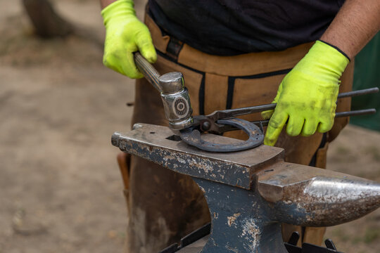 Blacksmith forging a horseshoe with a hammer onto anvil at the foreground. Forging furnace. Farrier fits hot horseshoe for horse. Changing a horseshoe. Blacksmith working with a horse. 