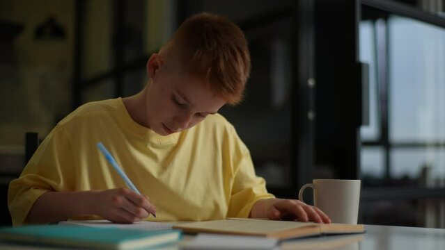 Cute red-haired pupil boy studying at home writing in exercise book doing homework, learning at home table. Smart schoolboy with freckles studying, reading textbook, flipping page of paper book.
