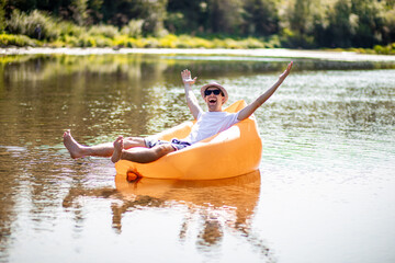 Cheerful caucasian good looking happy guy in sunglasses sitting on orange inflatable mattress with...