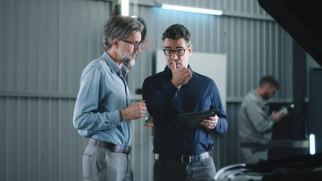 Dolly shot in repair shop of car service manager in glasses and casual suit with a tablet, talking to a man and discussing car diagnostics and repairing