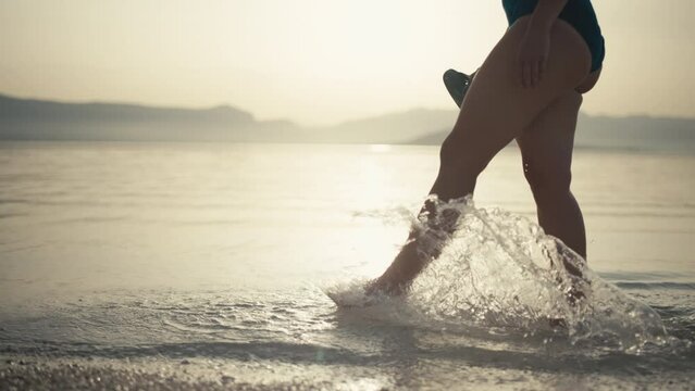 Slow motion video of a girl walking on the beach at sunrise wetting her feet in the ocean water. Close-up of female legs walking by the sea. High quality 4k footage