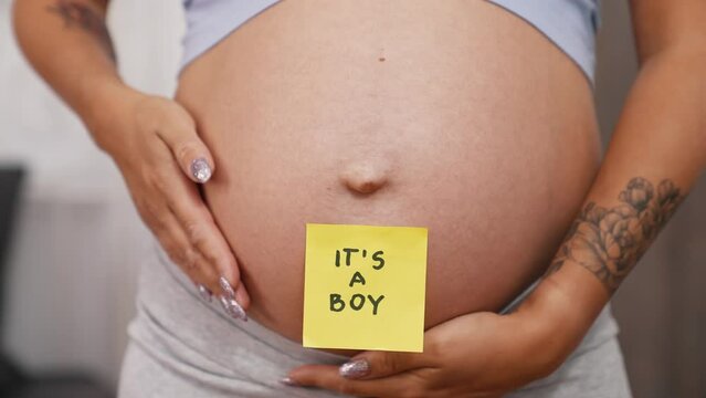 Gender reveal concept. It's a boy. Yellow sticker placed on a caucasian pregnant person's belly. Tattoo on arm, long nails, biker shorts. Middle body bart photo. High quality 4k footage