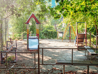 Small empty playground in the yard. A small children's fenced playground on sandy ground strewn with dry foliage, in the shade of green summer trees
