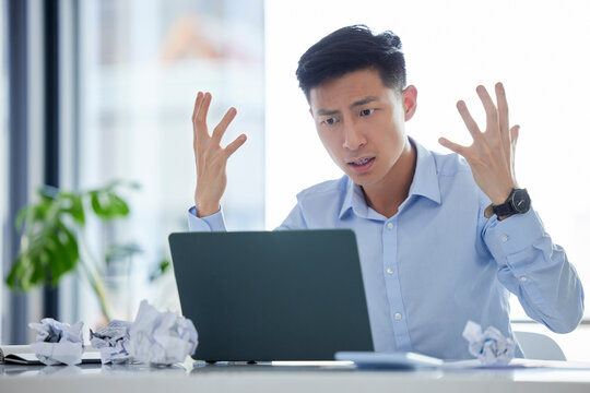 Frustrated, annoyed and irritated business man looking angry while working on his laptop. Young male worker feeling upset or struggling with an internet crash while at his desk in the office