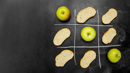Tic-tac-toe game between harmful calories and healthy food, apples and pieces of white bread on a black graphite board, tic-tac-toe game with white chalk, diet food problems