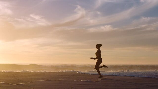 Silhouette of active, healthy and fit woman running on a beach at sunset in slow motion. Young focused and determined female exercising and training outside near the ocean at dawn dusk or sunrise