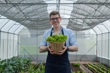 A Caucasian male farmer looks and gives basket of fresh vegetables to camera with a happy smile in plantation greenhouse. Gardener man collects natural organic produce from agriculture nursery crops.
