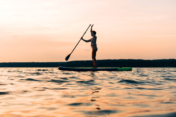 Joyful sport woman is training on a SUP board on a large lake during the evening. Stand up paddle...