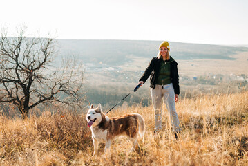 Young woman and her husky dog trek down a forest trail offering a scenic view of the trees changing...
