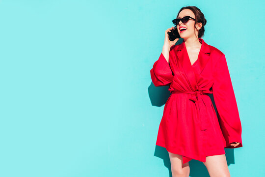 Young Beautiful Smiling Female In Trendy Summer Red Dress. Sexy Carefree Woman Posing Near Blue Wall In Studio. Positive Model Having Fun. Holding Smartphone. Talking At Phone With Friends