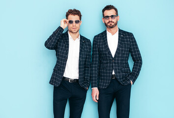 Portrait of two handsome confident stylish hipster lambersexual models. Sexy modern men dressed in black elegant suit. Fashion male posing in studio near blue wall. In sunglasses