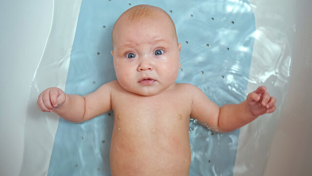 Baby girl lies in bathtub with water. Child looks around. Baby daughter likes to bathe waving hands with wonderment closeup