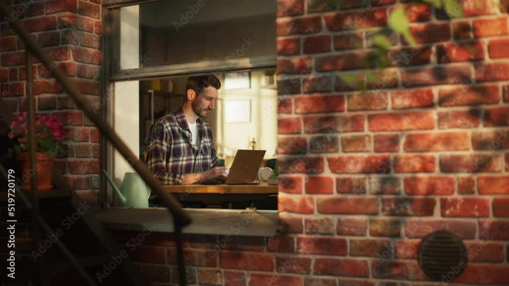 Poster Male Software Engineer Coding on Laptop Computer While Working in Modern Apartment. Professional IT Specialist Creating Application. Remote Working Freelancer. View From Outdoors into Window.