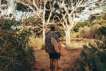 Surfer z deską surfingową, idący wybrzeżem na naturalnym tle, drzewa i kaktusy. © insomniafoto