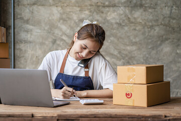 Young Asian Woman working from home, talking to clients with smartphone and working in happy mood with laptop and box. People with online shopping ideas, SME entrepreneurs or freelancers