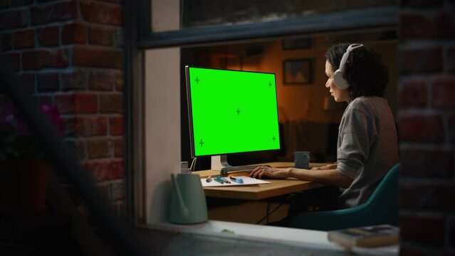 Female Designer Using Desktop Computer with Green Screen Chroma Key Monitor. Remote Access Manager Listening to Music or Podcast in Headphones, Freelancer Working at Night. Zoom Out, Window Shot.