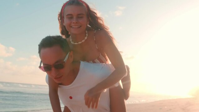 Young attractive romantic Caucasian couple date alone with each other having fun relaxing on beach man carries woman in swimsuit on back walking on sand washed by ocean. Love, relationships