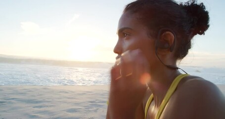 Fit, sporty and active female removing her earphones after a workout at the beach. Tired and exhausted woman resting and taking a break after exercise with waves in the sunset background