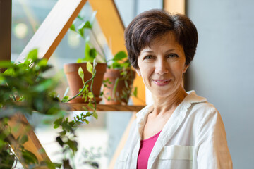 portrait of cute middle aged jewish woman in cozy green house. plant care. happy female smiling, care for flowers. beautiful stylish adult jew home gardening