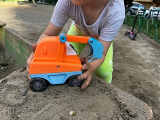 A kid takes their bright toy excavator with tiny hands in sand. Playing with toy truck in a sandbox on a playground