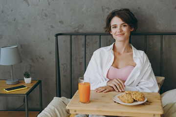 Young smiling happy woman wear white shirt pajama she lying in bed eat breakfast look camera rest relax spend time in bedroom lounge home in hotel room wake up dream be lost in reverie good mood day.