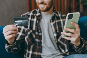 Cropped young man he wear brown shirt use mobile cell phone hold credit bank card shopping online order delivery booking tour sit on blue sofa in own living room apartment stay home indoor flat on