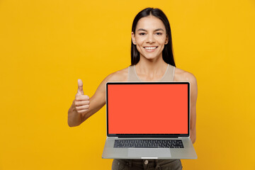 Young smiling fun happy satisfied latin woman 30s she wear basic beige tank shirt hold use work on laptop pc computer with blank screen workspace area show thumb up isolated on plain yellow backround