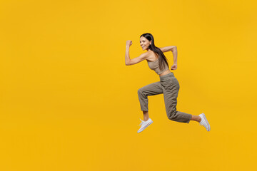 Full body side view smiling happy fun cool young latin woman 30s she wear beige tank shirt jump high run fast look camera isolated on plain yellow backround studio portrait. People lifestyle concept.