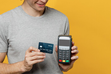 Cropped fun young man 20s he wear grey t-shirt hold wireless modern bank payment terminal to process acquire credit card payments isolated on plain yellow backround studio. People lifestyle concept.