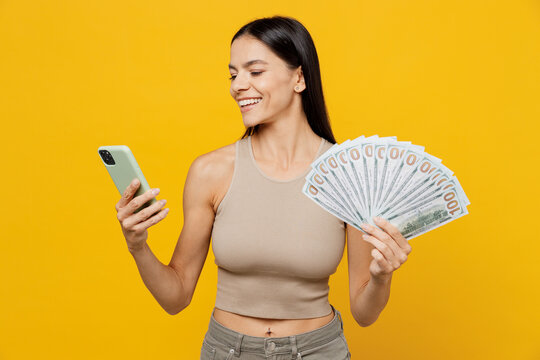 Young latin woman 30s she wear basic beige tank shirt holding fan of cash money in dollar banknotes use mobile cell phone isolated on plain yellow backround studio portrait. People lifestyle concept.