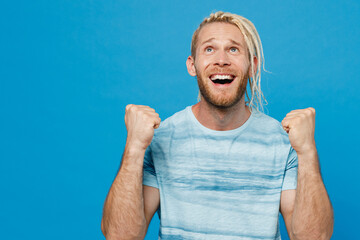 Young excited blond man with dreadlocks 20s he wear white t-shirt doing winner gesture celebrate clenching fists say yes isolated on plain pastel light blue background studio People lifestyle concept.