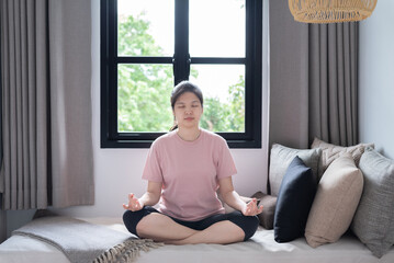 Cheerful young Asian woman in pink t-shirt and leggings sitting in Ardha Padmasana position on couch near windows, practicing yoga, doing half Lotus pose with mudra gesture. Yoga,well-being concept.