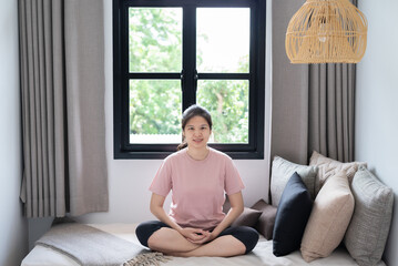 Cheerful young Asian woman in pink t-shirt and leggings sitting in Ardha Padmasana position on couch near windows, practicing meditation, smiling. Yoga, well-being, mindfulness lifestyle concept.