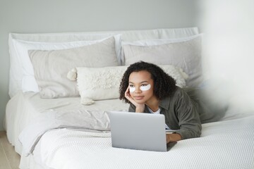 Weekend in bed. Lovely afro girl applies eye patches and relaxing at home watching movie.