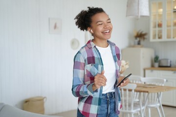 Excited happy girl in airpods is holding mobile phone. Teenage girl is singing in weekend morning.