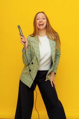 Vertical shot of joyful pretty woman singing a song with a microphone, presenting an event or having a party, enjoy the moment, standing over yellow background in casual clothes. Young emotional woman
