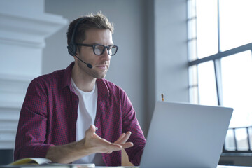 European man online tutor wearing glasses sitting at desk in headset and talking by video call