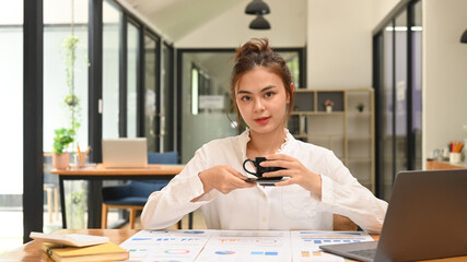 Portrait of young female employee holding coffee cp and looking confidently to camera