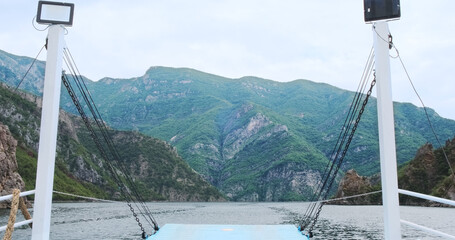 Back view of ferry crossing mountain Komani lake in summer Albania. Beautiful mountains covered green plants, trees. Ship moves on turquoise lake water. Ferry travel by river. Beauty boat trip