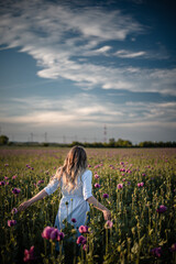 Young woman with long blonde hair is walking in a beautiful field of violet poppy flowers