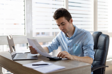 Young businessman checking bills, holding receipt, using calculator, accountant calculating company expenses, financial report, sitting at wooden work desk, young male planning budget