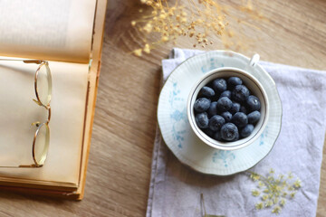 Vintage porcelain cup filled with fresh blueberries, pressed flowers, open book, reading glasses and lit candle on the table. Hygge at home. Top view.