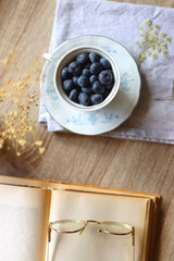 Vintage porcelain cup filled with fresh blueberries, pressed flowers, open book, reading glasses and lit candle on the table. Hygge at home. Top view.