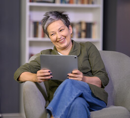 Happy smiling middle age grey-haired woman makes emotional online conversation over digital tablet sitting in chair at home. Elderly and technology concept.