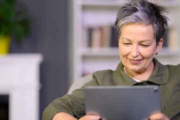 Happy mature grey-haired woman reading news looking into own pad gadget in hand. Modern smiling...