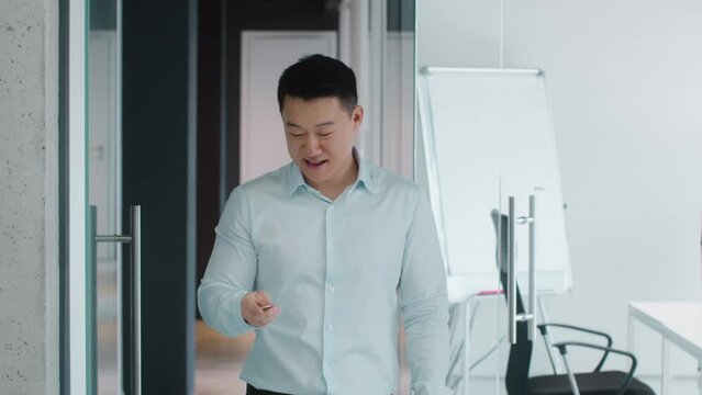 Positive middle aged asian businessman playing with btc coin, showing bitcoin to camera, standing at office, free space