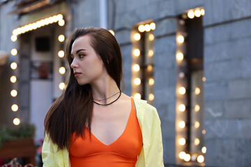 Portrait of pretty young girl with long hair and makeup wearing orange top on a street on blurred lights background. Female fashion, romantic evening, holiday in city