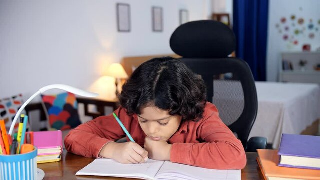 A young boy with black curly hair is completing his homework sitting at a desk - school assignments  learning and knowledge. A little boy in casual clothes is studying alone at home - a studious ch...