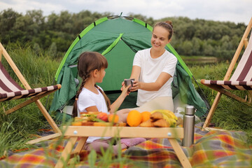 Naklejka na ściany i meble Image of charming delighted young adult woman and little girl sitting on blanket near tent having picnic, daughter giving a cup of tea or coffee to her mommy.