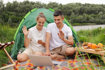 Naklejka na ściany i meble Full length portrait of happy couple having rest near river, staying overnight outdoor, enjoying nature, having video call with friends, waving hands, saying hello or bye.